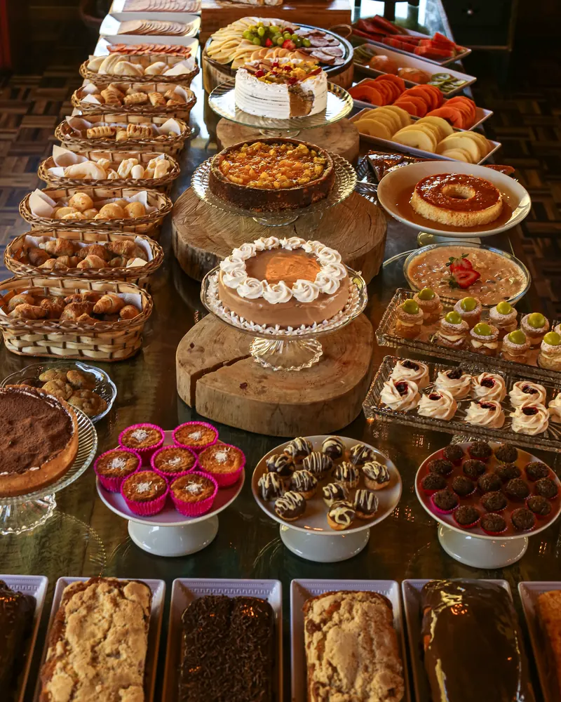 Mesa farta do café colonial com bolos, tortas, pudim, docinhos e pães.