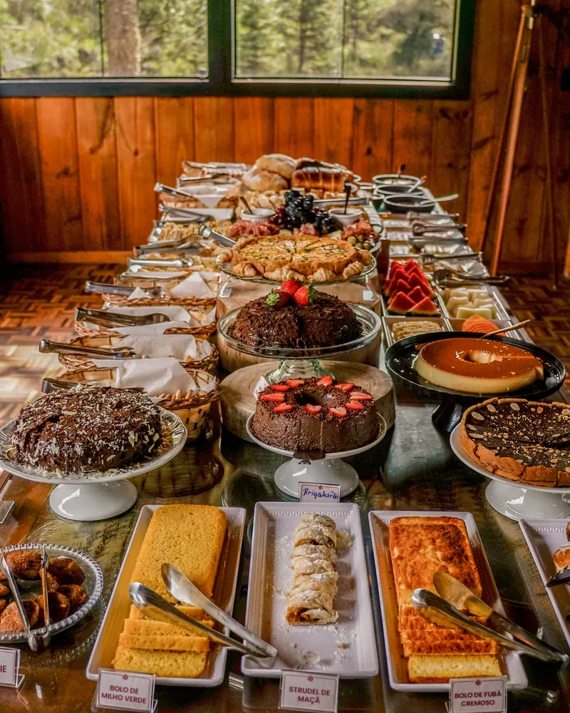 Mesa do café colonial junto às janelas, com bolos, tortas e doces.
